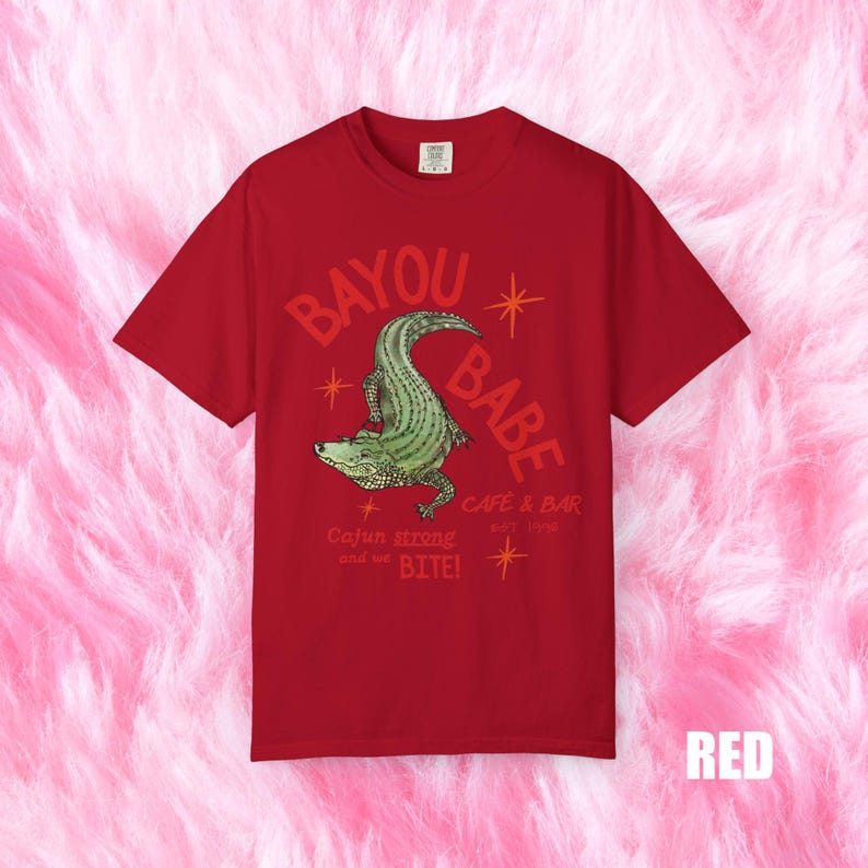 Op de afbeelding: Rood T-shirt met de tekst "BAYOU BABE" en een groene alligatorafbeelding. Het shirt bevat ook de tekst "Cajun strong and we BITE!" en "CAFE & BAR EST. 1996".