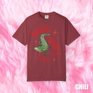 Op de afbeelding: Chili-kleurig T-shirt met een groene alligator illustratie en de rode tekst "BAYOU BABE". Onder de alligator staat de tekst "Cajun Kitchen" en "Bite!". Het shirt heeft een losse pasvorm.