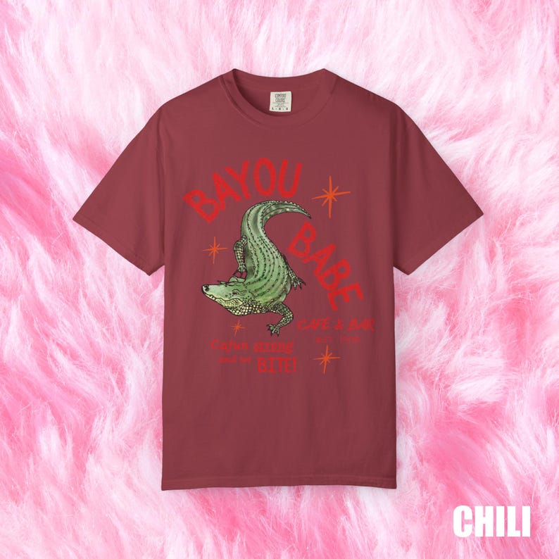 Op de afbeelding: Chili-kleurig T-shirt met een groene alligator illustratie en de rode tekst "BAYOU BABE". Onder de alligator staat de tekst "Cajun Kitchen" en "Bite!". Het shirt heeft een losse pasvorm.