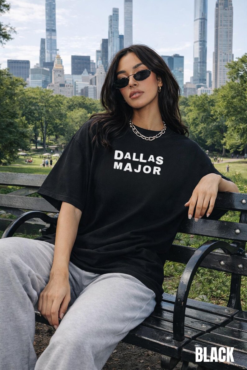 May include: Black t-shirt with the text "DALLAS MAJOR" in white block letters. The model is wearing sunglasses and a silver chain necklace. The word "BLACK" is at the bottom right corner of the image.