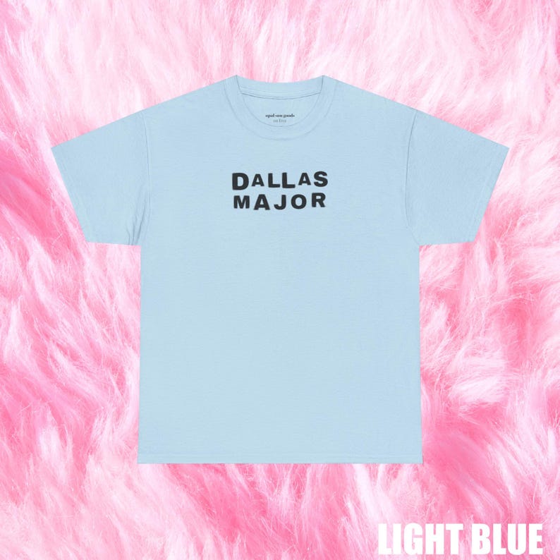 May include: Light blue t-shirt with the text "DALLAS MAJOR" in black block letters. The shirt is a light blue color and has a crew neck. The words "aged are goods" are printed on the top of the shirt.