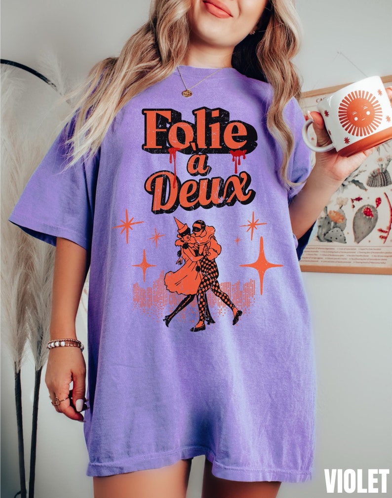 May include: Purple t-shirt with a vintage-style graphic of two people dancing. The graphic says "Folie a Deux" in orange and red lettering.