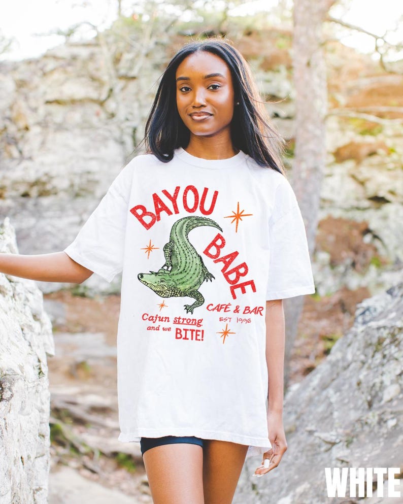 Op de afbeelding: Wit T-shirt met de tekst "BAYOU BABE" in het rood, met een groene alligatorafbeelding. Het shirt bevat ook de tekst "CAFE & BAR EST 1996" en "Cajun strong and we BITE!"