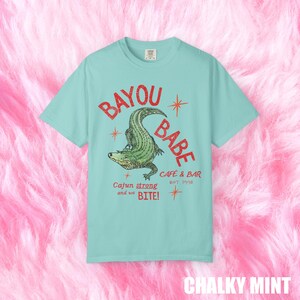 Op de afbeelding: Mintgroen t-shirt met een distressed graphic van "BAYOU BABE" en een alligator. De tekst "Cajun strong and we BITE!" en "CAFE & BAR EST 1996" staan ook op het shirt.