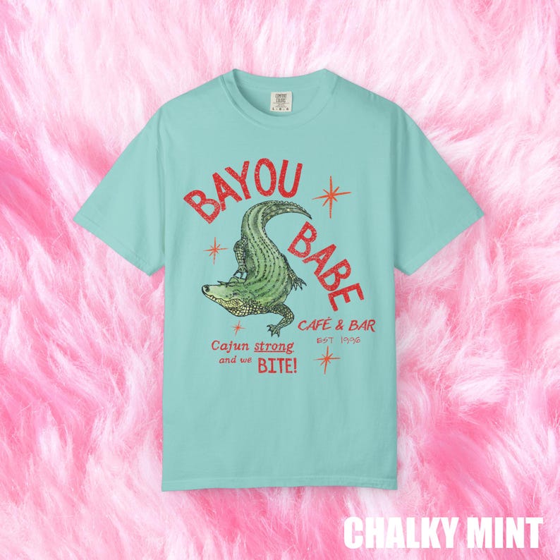 Op de afbeelding: Mintgroen t-shirt met een distressed graphic van "BAYOU BABE" en een alligator. De tekst "Cajun strong and we BITE!" en "CAFE & BAR EST 1996" staan ook op het shirt.