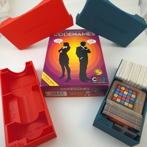 May include: A red and blue game box set for the board game Codenames. The box set includes a game board, word cards, key cards, agent cards, and a code word card. The game is a word association game where players try to guess the secret words of their team.