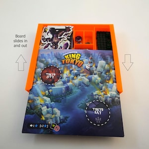 以下が含まれることがあります： キング・オブ・トーキョーボードゲーム用のスライド式ボードが付いたオレンジ色のプラスチック製ゲームオーガナイザー。オーガナイザーには、ゲームピース、カード、サイコロ用のコンパートメントがあります。背景にはボードゲームが見えます。
