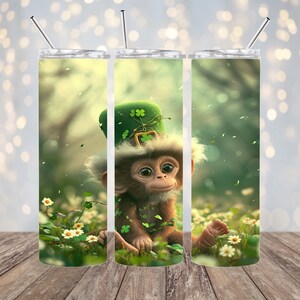 Può includere: Un bicchiere in acciaio inossidabile con uno sfondo verde che presenta una scimmia dei cartoni animati che indossa un cappello da folletto verde con una fibbia dorata. La scimmia è circondata da trifogli verdi e margherite bianche.