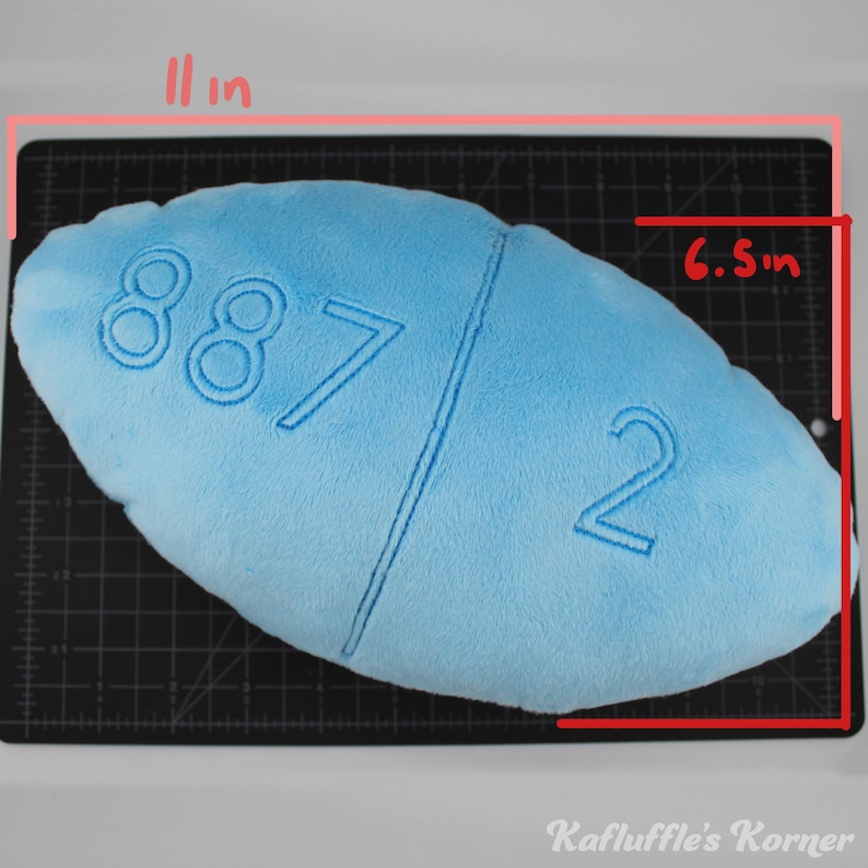 May include: A light blue plush pill-shaped pillow, measuring 28 cm long and 16.5 cm wide. The pillow features embroidered numbers "887" and "2". The text "Kafluffle's Korner" is visible at the bottom of the image.