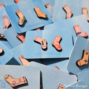 Può includere: Una collezione di carte quadrate azzurre con abbellimenti in legno a forma di calzino. I calzini sono in vari colori, tra cui rosa, arancione e verde. Il testo "KAFLUFFLE" è visibile su alcune carte.