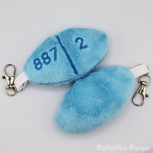 May include: Two light blue plush pill-shaped keyrings with the numbers "887" and "2" embroidered on one. Each keyring has a white strap and a silver-coloured clasp. The keyrings are on a white surface.