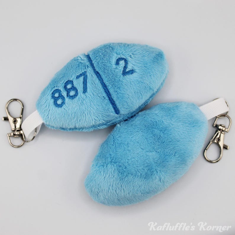 May include: Two light blue plush pill-shaped keyrings with the numbers "887" and "2" embroidered on one. Each keyring has a white strap and a silver-coloured clasp. The keyrings are on a white surface.