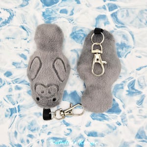 May include: Two gray plush manatee keychains with embroidered details. One keychain has a silver-colored clasp and a black strap. The other manatee keychain is without a clasp. The background is a blue and white pattern.