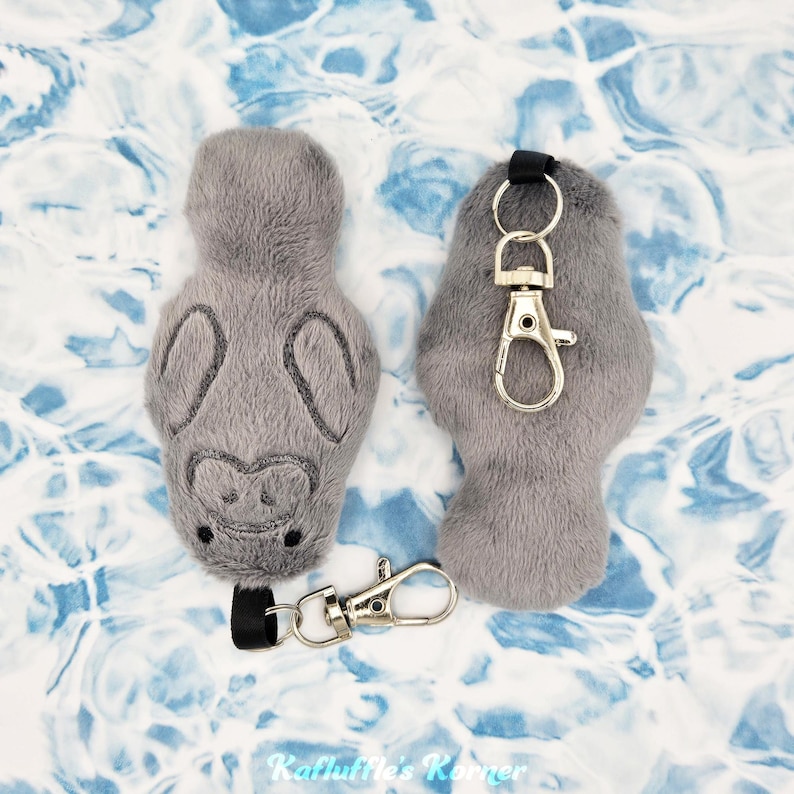 May include: Two gray plush manatee keychains with embroidered details. One keychain has a silver-colored clasp and a black strap. The other manatee keychain is without a clasp. The background is a blue and white pattern.