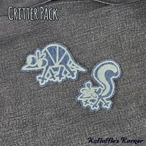 May include: Two denim fabric patches featuring embroidered outlines of an armadillo and a skunk. The text CRITTER PACK is printed at the top, and Kafluffle's Korner is printed at the bottom. The patches are displayed on a dark grey denim background.