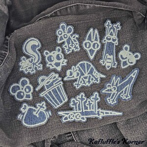 May include: A collection of twelve embroidered patches featuring cartoon-style characters and objects, including a skunk, a trash can, and a sword, stitched in light blue thread onto a dark grey denim fabric background with the text Kafluffle's Korner.