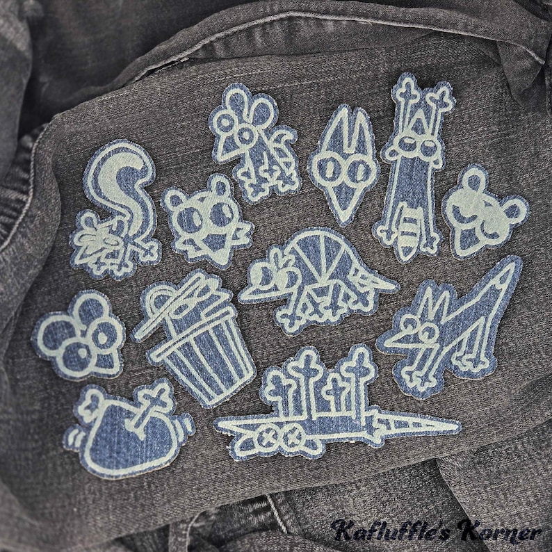 May include: A collection of twelve embroidered patches featuring cartoon-style characters and objects, including a skunk, a trash can, and a sword, stitched in light blue thread onto a dark grey denim fabric background with the text Kafluffle's Korner.