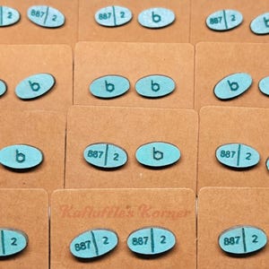 May include: A collection of teal-colored, oval-shaped wooden earrings. Some earrings have the number "887 2" printed on them, while others have the letter "b". The earrings are displayed on brown cardboard cards. The text "Kafluffle's Korner" is visible.