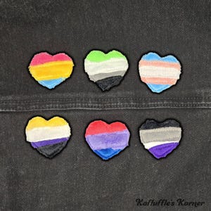 May include: Six heart-shaped patches with various pride flag designs. The patches feature colors representing pansexual, asexual, transgender, non-binary, bisexual, and aromantic identities. The patches are on a dark denim background.