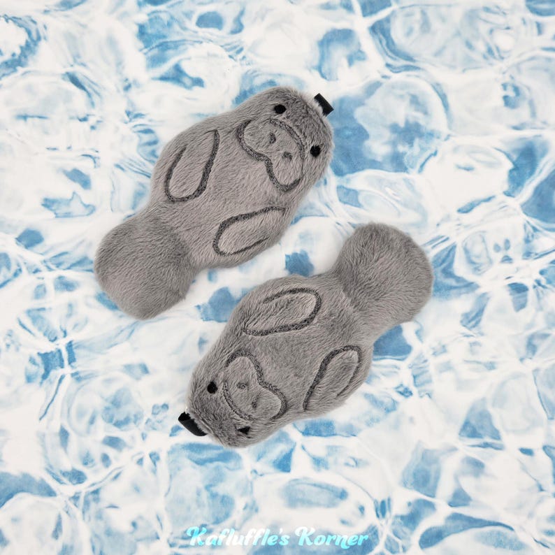 May include: Two gray plush manatee toys with black accents, lying on a blue and white water-like background. The manatees have embroidered details and a small black tag. The toys are designed to resemble the sea creature.