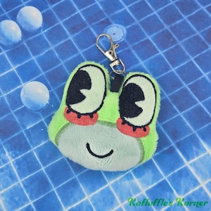 May include: A plush frog keychain featuring a light green fuzzy texture, large embroidered black eyes with white highlights, red accents under the eyes, and a simple stitched smile. A silver metal lobster clasp is attached to the top.