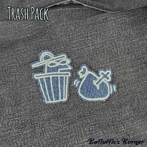 May include: Two denim fabric patches featuring light-coloured embroidered outlines. One patch depicts a trash can with a lid, and the other shows a trash bag with a bone. The text TRASH PACK appears at the top, and Kafluffle's Korner at the bottom.