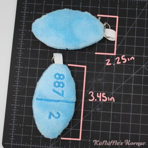 May include: Two light blue plush keyrings. One is oval-shaped, measuring 5.7 cm long. The other is pill-shaped, 8.8 cm long, with the number "887 2" embroidered on it. Both have a metal ring and a white fabric loop.