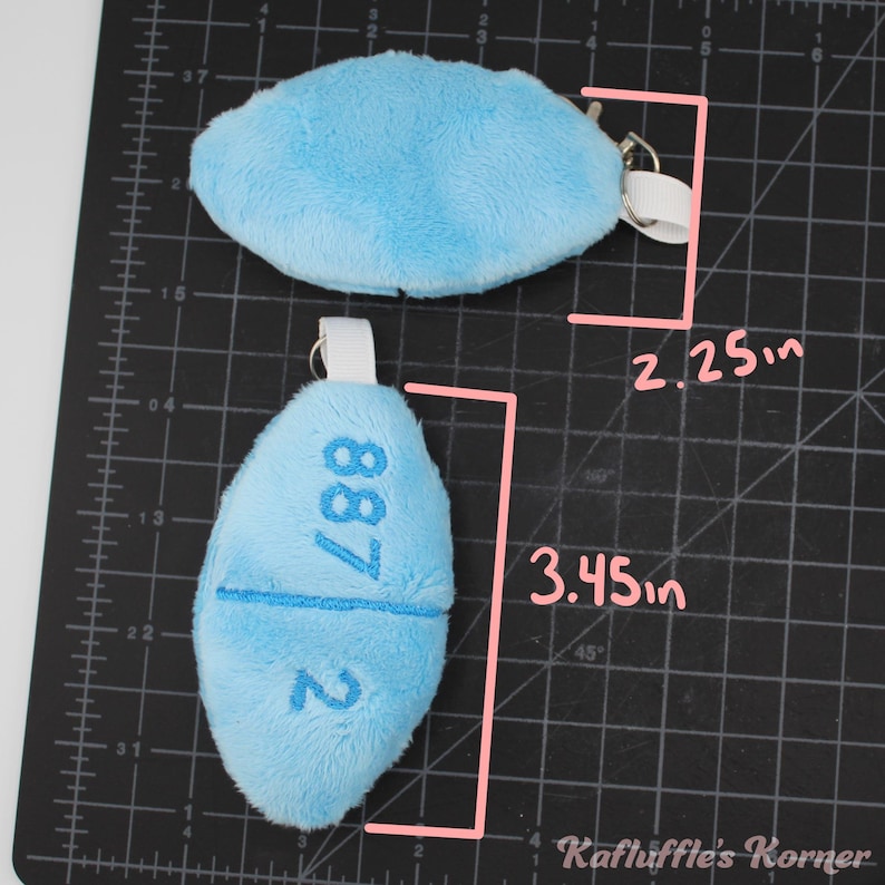 May include: Two light blue plush keyrings. One is oval-shaped, measuring 5.7 cm long. The other is pill-shaped, 8.8 cm long, with the number "887 2" embroidered on it. Both have a metal ring and a white fabric loop.