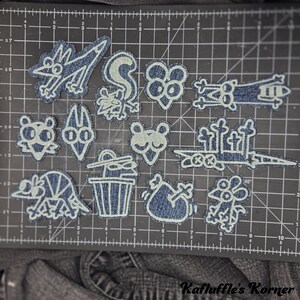 May include: A collection of twelve denim fabric patches featuring white embroidered line art of various cartoon characters and objects, including animals, a trash can, and a graveyard scene, arranged on a black cutting mat with centimetre markings.