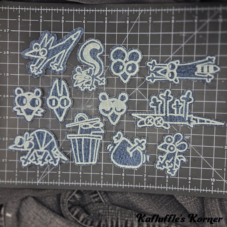 May include: A collection of twelve denim fabric patches featuring white embroidered line art of various cartoon characters and objects, including animals, a trash can, and a graveyard scene, arranged on a black cutting mat with centimetre markings.