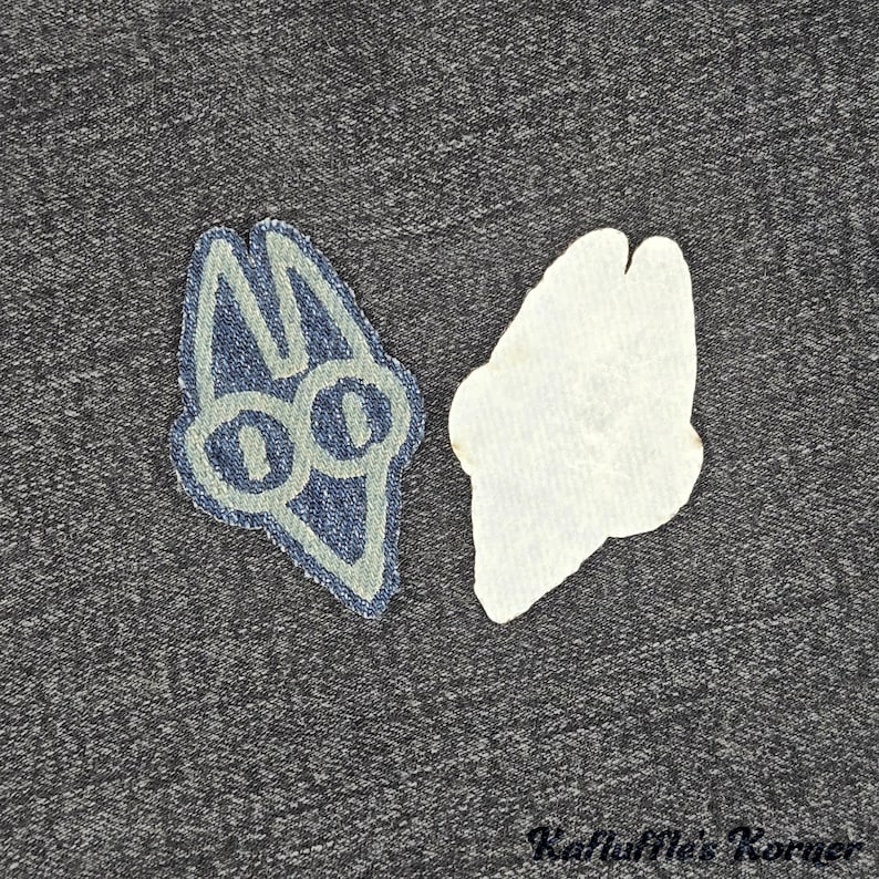 May include: A denim fabric patch featuring a stylised, light blue embroidered outline of a cat-like head with two large circular eyes and pointed ears, shown alongside its plain white adhesive backing on a dark grey textured fabric surface.