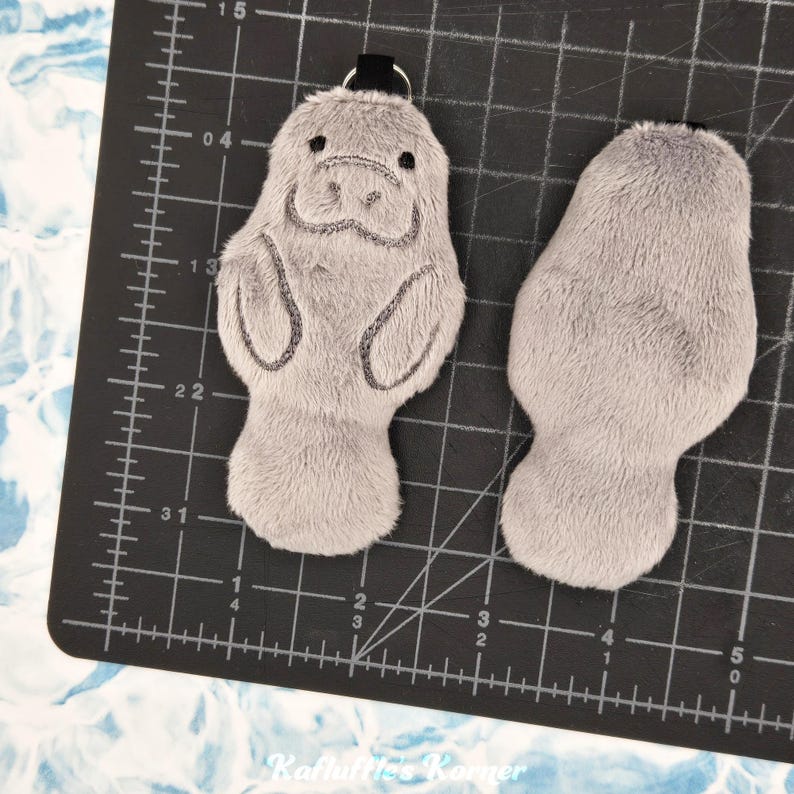 May include: Two plush manatee keychains. One keychain is facing forward, showing the manatee's face, with black embroidered details. The other keychain is the back of the manatee. Both are gray and have a black loop for attaching to keys.