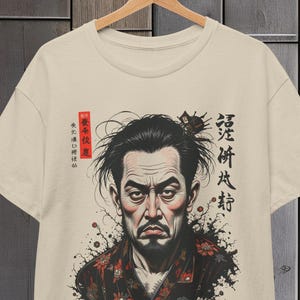 Op de afbeelding: Beige t-shirt met een gedetailleerde illustratie van een ernstig kijkende samoerai. Het kunstwerk bevat Japanse kalligrafie en een rood zegel. De samoerai draagt een kledingstuk met patroon en het ontwerp heeft een vintage esthetiek.
