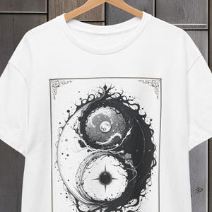 Puede incluir: Camiseta blanca con un diseño Yin Yang en blanco y negro. El gráfico muestra un patrón arremolinado con detalles intrincados, enmarcado por un borde decorativo. La camiseta está colgada en una percha de madera sobre un fondo de madera.