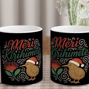 May include: White ceramic mug with a black background featuring the text "Meri Kirihimete" in red and green script. A cartoon kiwi bird wearing a Santa hat is next to a red flower. Decorative swirls complete the design.