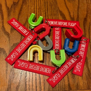 May include: A collection of colorful keychains with the text "REMOVE BEFORE FLIGHT" printed on red fabric tags. The keychains are shaped like a U and are made of different colors, including green, blue, yellow, and gray.