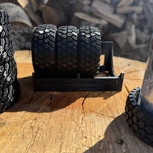 May include: A 3D printed black tire rack with four black tire-shaped holders. The rack is sitting on a wooden surface. The text "SoCal3DPrintsDesigns Etsy.com" is printed on the front of the rack.