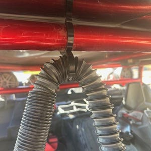 May include: A black flexible hose is secured to a red metal bar with a black plastic zip tie. The hose is coiled and appears to be made of a durable material.