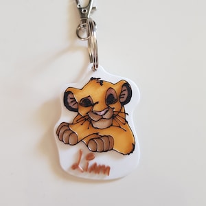 Könnte beinhalten: Weißer Schlüsselanhänger mit einem Cartoon-Bild von Simba aus dem König der Löwen. Der Schlüsselanhänger hat einen silbernen Metallring und einen Karabiner. Der Name "Liam" ist in braunen Buchstaben unter dem Bild geschrieben.