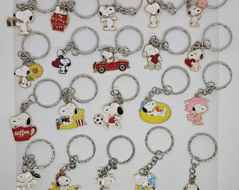 May include: A collection of twenty metal keychains featuring Snoopy and Woodstock in various poses, including holding a gift, driving a red car, playing soccer, sitting in a coffee mug, and wearing costumes, all attached to silver split rings.