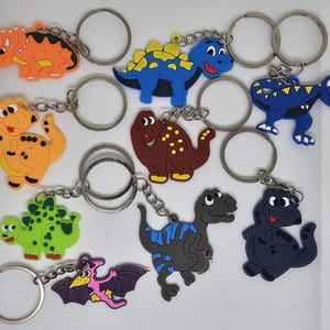 Könnte beinhalten: Ein Set mit zehn bunten Dinosaurier-Schlüsselanhängern. Jeder Schlüsselanhänger zeigt ein anderes Dinosaurier-Design, darunter ein blauer Stegosaurus, ein grüner Triceratops, ein brauner Brachiosaurus, ein roter Velociraptor, ein lila Pterodactylus, ein gelber Tyrannosaurus Rex, ein orangefarbener Ankylosaurus, ein schwarzer Parasaurolophus, ein rosa Spinosaurus und ein weißer Carnotaurus. Die Schlüsselanhänger sind aus weichem, strapazierfähigem Kunststoff gefertigt und perfekt, um Ihren Schlüsseln, Taschen oder Rucksäcken eine lustige Note zu verleihen.