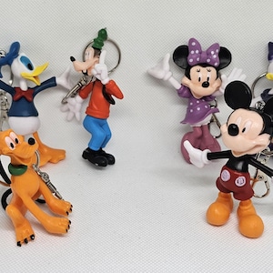 Peut inclure: Ensemble de porte-clés de personnages Disney, dont Mickey Mouse, Minnie Mouse, Donald Duck, Daisy Duck, Dingo et Pluto. Chaque figurine est de couleur vive et attachée à un porte-clés argenté. Les personnages sont dans diverses poses.