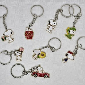 Może przedstawiać: Kolekcja breloków do kluczy z postaciami Snoopy'ego. Każdy brelok przedstawia inny wzór Snoopy'ego, w tym Snoopy z prezentem, w samochodzie i w kapeluszu. Breloki mają srebrne łańcuszki i kółka.