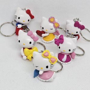 May include: Six white Hello Kitty keychains with different colored outfits and accessories. The keychains feature Hello Kitty in various poses, including holding a heart, playing a violin, and wearing angel wings.