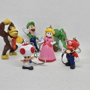 Könnte beinhalten: Eine Sammlung farbenfroher Super Mario Bros. Charakter-Schlüsselanhänger. Die Schlüsselanhänger zeigen Charaktere wie Mario, Luigi, Prinzessin Peach, Yoshi, Donkey Kong und Toad. Jede Figur ist detailliert und an einem silbernen Schlüsselanhängerring befestigt.