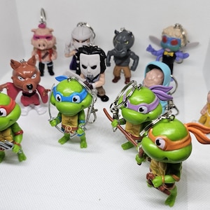 May include: A collection of 12 colourful plastic keychains featuring various characters, including Teenage Mutant Ninja Turtles, a pig, a wolf, and a rhino. The keychains are all attached to metal rings.