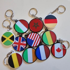 Puede incluir: Una colección de 11 llaveros redondos con diferentes banderas de países impresas en ellos. Los llaveros son blancos con bordes negros y presentan banderas de Jamaica, Reino Unido, Estados Unidos, Marruecos, Alemania, Irlanda, Francia, Canadá, Bélgica, Italia y Senegal.