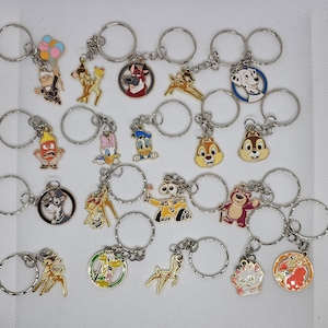 Könnte beinhalten: Sammlung von Charakter-Schlüsselanhängern mit silbernen Metallringen. Die Schlüsselanhänger zeigen verschiedene Zeichentrickfiguren in leuchtenden Farben, darunter einen Dalmatiner, Chip und Chap und Donald Duck. Jeder Schlüsselanhänger ist ein kleiner, detaillierter Anhänger.
