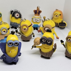 May include: A collection of ten yellow Minion keychains. Each keychain features a different Minion character with unique clothing and accessories. The Minions are wearing various outfits, including a blue and purple cape, a brown coat, a king's crown, and a brown fur coat.