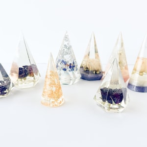 May include: A selection of clear resin ring holders in geometric shapes. Each holder contains dried flowers, gold leaf, or other decorative elements. The holders are displayed on a white surface, highlighting their individual designs.
